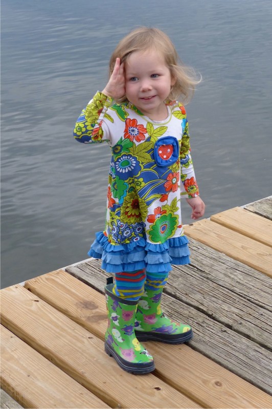 two-year old girl in colorful outfit with green rubber boots standing on pier.