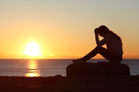 Silhouette of woman with head in hands sitting on rock at shore at sunrise