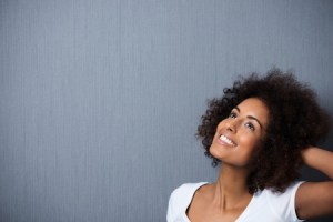 25363197 - beautiful young african american woman standing daydreaming with her hand to her curly afro hair and a whimsical smile of pleasure as she looks up into the air, with copyspace