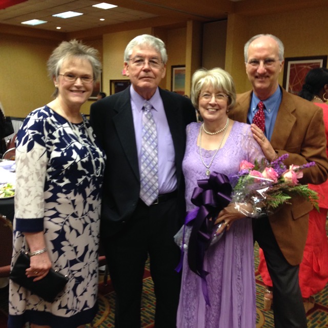 A group of four people smiling at a celebration, with one person holding a bouquet of flowers.