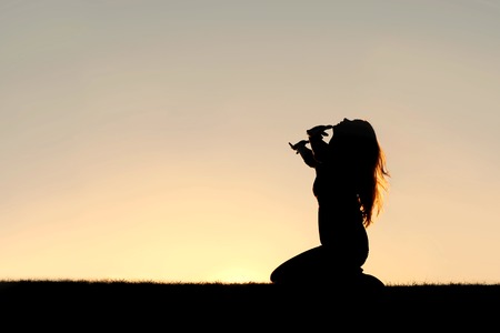 a silhouette of a woman kneeling down with her hands in the air, praying, thanking, and surrendering to god