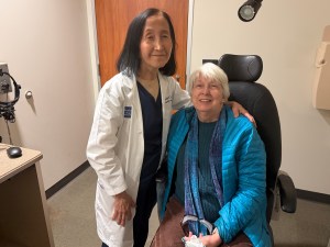 A smiling healthcare professional in a white coat stands next to a patient sitting in a chair, both are in a medical office.