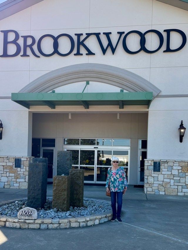 A woman stands in front of the Brookwood building, featuring a stone sculpture and a sign displaying the name 'Brookwood'.